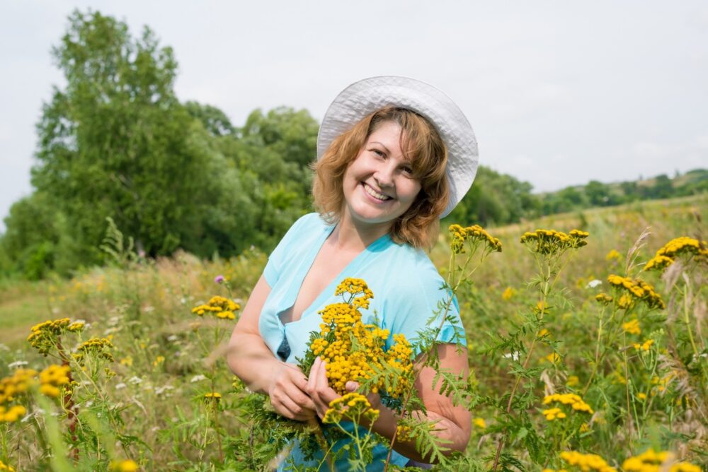 A smiling woman in a hat is gathering herbs in a meadow. She is holding a bouquet of herbs in her hand.