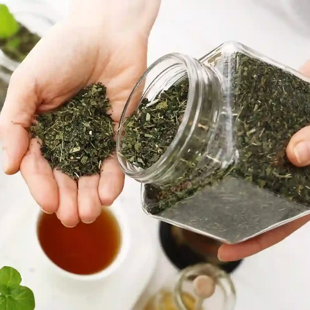 Hands holding a glass jar of dried green herbs, pouring some into an open palm.