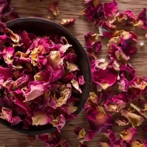 Top view of dried red rose petals in a bowl spilling onto wooden table by Health Embassy